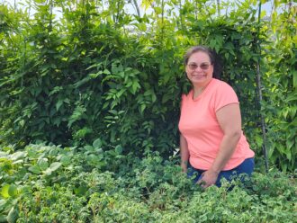 a farmer poses. in the background are hopniss (American groundnut) plants. Oca is in the foreground to the viewer's right and on the left are African potato mint plants.