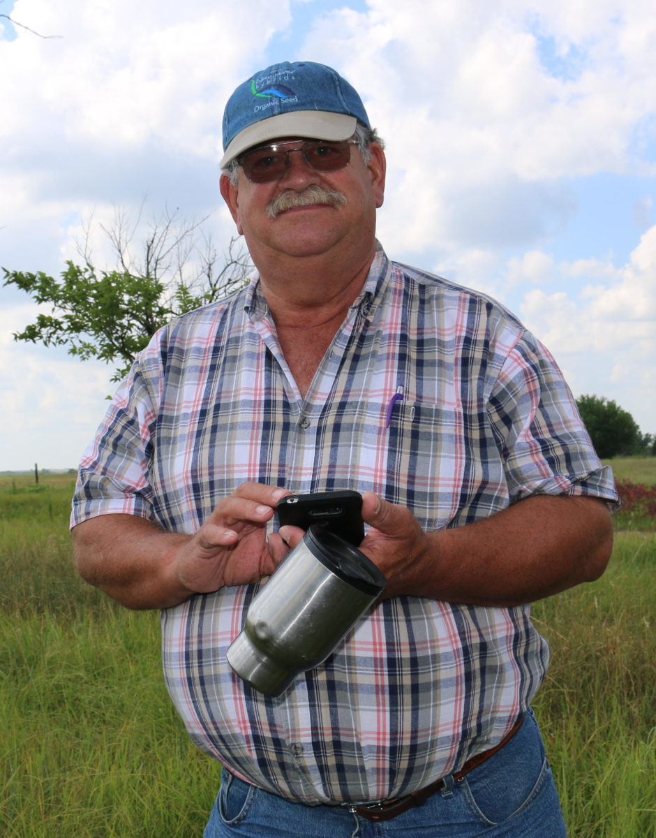 Donn Teske holding his phone and coffee cup in a field wearing a hat and sunglasses