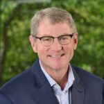 man in navy blazer, with glasses in front of green leaves
